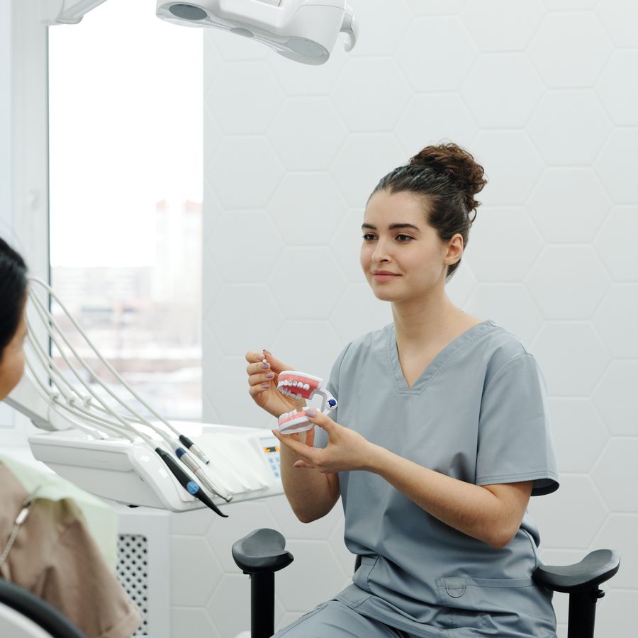 Dentista sentada ao lado da cadeira odontológica explica um modelo de arcada dentária para uma paciente